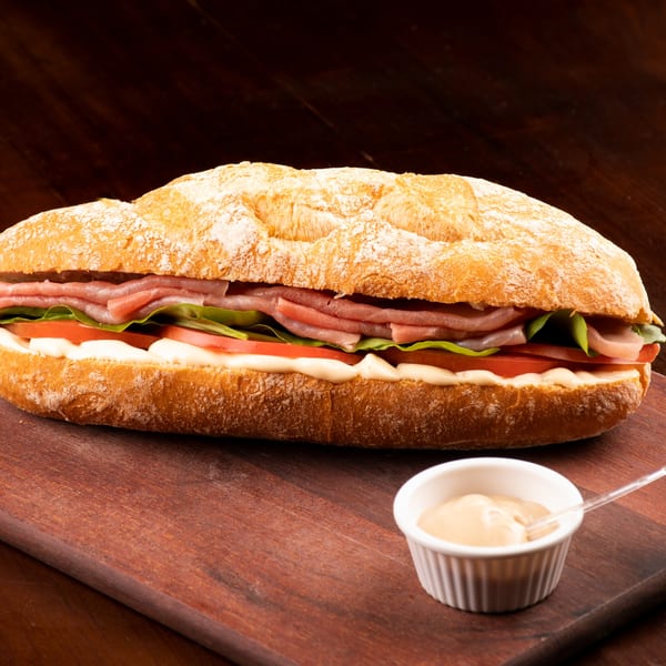 A large baguette sandwich with ham, lettuce, tomatoes, and mayonnaise, served with a side of sauce.