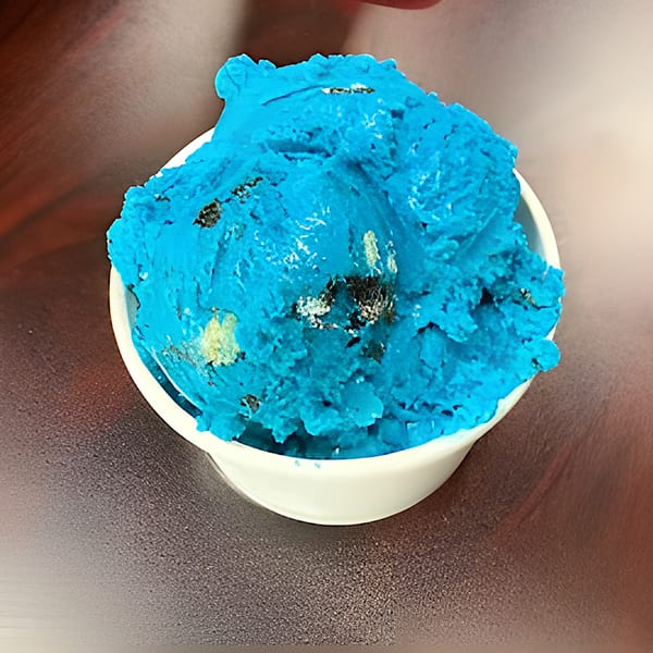 Bright blue ice cream with dark cookie chunks in a white cup on a dark surface.